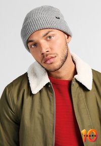 Szara, prążkowana czapka beanie noszona z oliwkową kurtką z białym, futrzanym kołnierzem na czerwonym, textured swetrze.