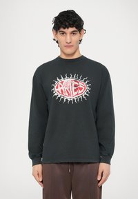 Tricou negru cu mânecă lungă, cu un grafic „Berbec” roșu și alb într-un oval înconjurat de un design cu raze de soare. Realizat din material de bumbac.