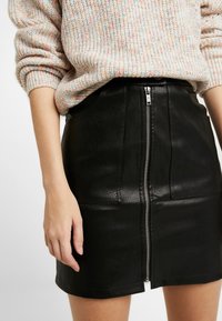 Black leather mini skirt with a front zipper closure, featuring a textured surface and clean lines. Worn with a textured, multicolored sweater.