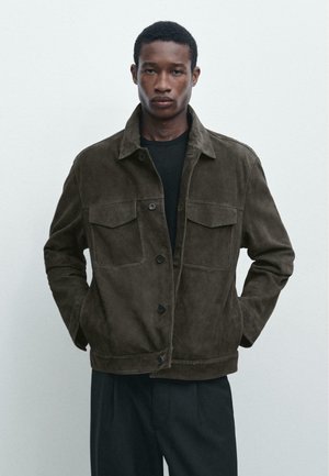 Junger Mann trägt dunkelolivgrüne, geknöpfte Wildlederjacke mit Klappentaschen auf der Brust und schwarze Hose, steht vor einfarbig hellem Hintergrund.