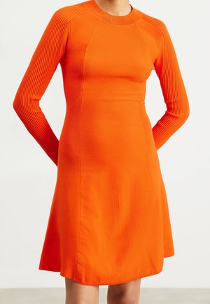 Mujer que lleva un vestido de punto de manga larga de color naranja brillante con mangas acanaladas y cuello redondo, de pie con las manos detrás de la espalda.