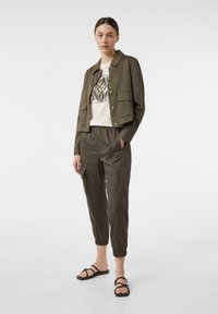 Oliwkowa kurtka z dwoma przednimi kieszeniami, zestawiona z odpowiednimi spodniami na gumce w tym samym kolorze oraz kremową koszulą z czarnym wzorem graficznym. Czarne sandały.