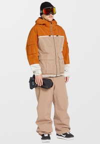 Snöjacka i orange och beige med dragkedjefickor och kontrasterande ärmar; ihop med beige byxor och svarta snöhandskar med logotyp.