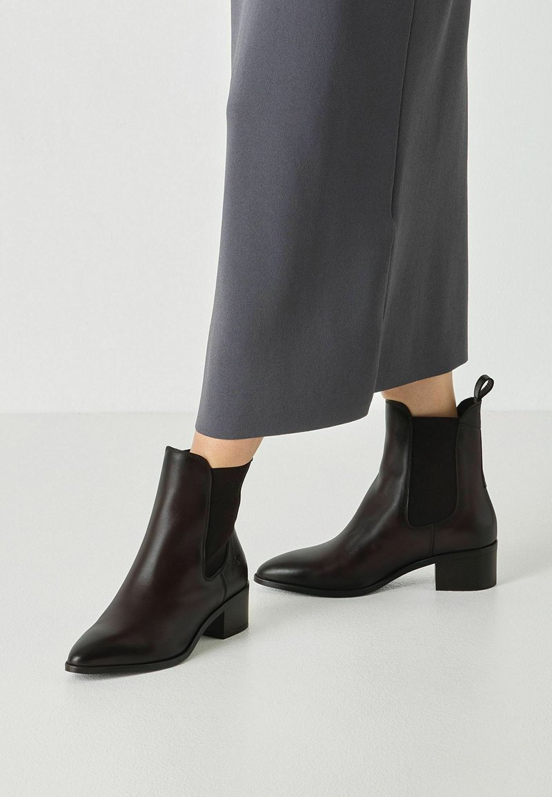 Bottes en cuir noires montantes jusqu’à la cheville avec des talons blocs bas, portées avec une longue jupe grise sur un fond clair uni.
