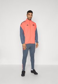 Adidas trainingspak in koraal en donkergrijs, met een rits aan de voorkant, logo-detail en gestreepte accenten op de mouwen en broekspijpen. Draagt met zwarte schoenen.