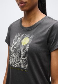 Γυναίκα που φοράει σκουρόγριφη μπλούζα με λευκά κλαδιά δέντρου και γραφικό κίτρινο ήλιο, με το κείμενο «Cumbia Sportswear» από κάτω.