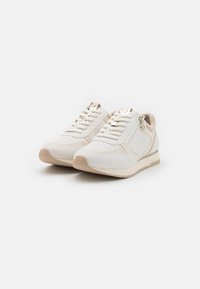 Baskets blanches avec des accents texturés, des lacets crème et une fermeture éclair sur le côté. Elles possèdent une semelle beige et des détails en métal doré.