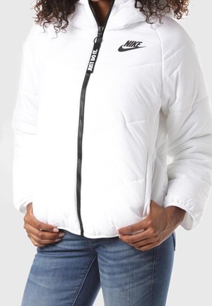 Weiße Nike Steppjacke mit schwarzem Reißverschluss und „Just Do It.“-Etikett, getragen zu blauen Jeans, Hände ruhen in der Nähe der Taschen, lockiges Haar sichtbar.