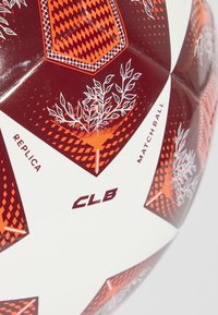 Baltas ir raudonas futbolo kamuolys su geometriniais raštais, oranžinėmis akcentais ir botaninių iliustracijų elementais. Žymėtas "REPLICA" ir "MATCH BALL" su "CLB."