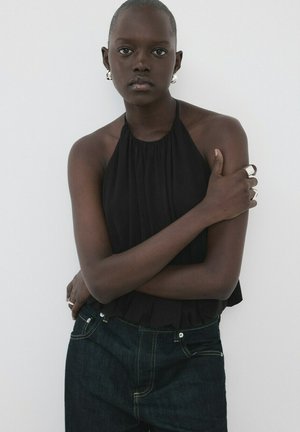 Femme aux cheveux courts portant un haut dos nu noir, un jean foncé, des boucles d'oreilles argentées et des bagues, debout les bras croisés contre un mur blanc uni.