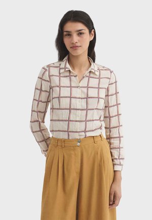 Frau mit langen dunklen Haaren, die ein beiges kariertes Hemd mit Knöpfen und senfgelbe, hochtaillierte, weit geschnittene Hosen trägt.