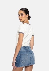 Młoda kobieta z ciemnymi włosami upiętymi w kok, ubrana w białą koszulę z krótkim rękawem z szarymi akcentami i niebieską dżinsową spódnicę, spoglądająca przez ramię.