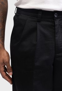Calças pretas plissadas com uma textura suave, apresentando um fecho de botão e laços para cinto, mostradas ao lado de uma mão com tatuagens visíveis.