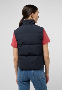 Gilet imbottito navy con colletto alto, caratterizzato da sezioni cucite orizzontalmente e una texture liscia, abbinato a una camicia rossa e jeans blu.