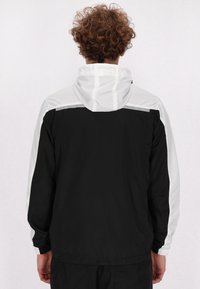 Sort og hvid vindjakke med hætte, elastiske manchetter og en let, glat tekstur. Bagtil ses farveblokdesign.