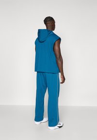 Tenue de sport pour homme en tissu teal avec un haut à capuche sans manches et un pantalon décontracté avec des rayures sur les côtés. Les chaussures sont blanches avec des accents noirs.