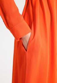 Main d'une personne insérée dans la poche latérale d'un vêtement à manches longues orange vif avec un poignet boutonné et un tissu lisse.