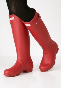 Bottes de pluie rouges en caoutchouc avec une texture lisse, une forme courbée et une semelle épaisse. Présentent une boucle sur le côté et une patch logo près du haut.
