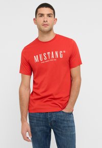 Czerwona bawełniana koszulka z krótkim rękawem i okrągłym dekoltem, z białym logo "MUSTANG" i napisem "JEANS COMPANY SINCE 1932" poniżej.