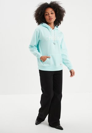 Lichtblauwe hoodie met een front pocket, witte tekst met "CREEK'S" en "Original Since 1974," gecombineerd met zwarte wijde broek en puntige schoenen.