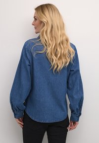 Camicia in denim blu con maniche lunghe leggermente gonfie, orlo arrotondato e dettagli con polsini bottonati, mostrata di spalle su uno sfondo neutro.