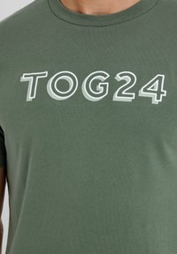 Green cotton t-shirt featuring white 3D text "TOG24." Classic crew neck design with short sleeves and a smooth, untextured surface.