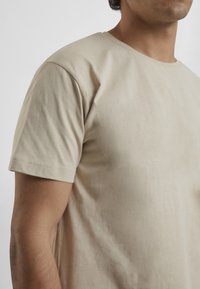 Beige színű pamut póló klasszikus kerek nyakkal és rövid ujjakkal, sima textúrával és minimális varrási részletekkel.