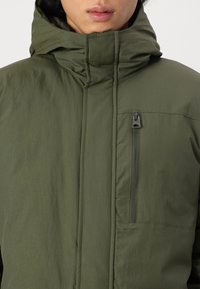 Vihreä hupullinen takki, jossa on sileä pinta, etutasku vetoketjulla ja näkyvät ompelusaumat. Mallissa on korkea kaulus ja minimalistinen tyyli.