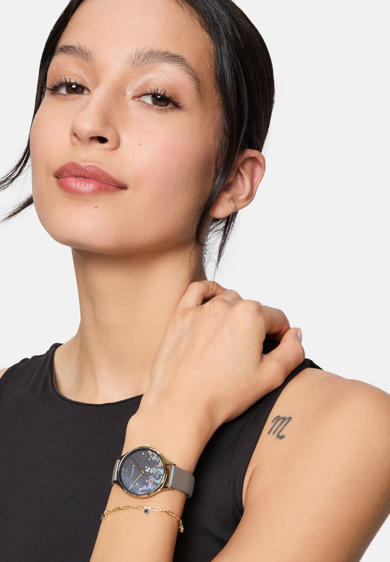 Graue Silikonarmbanduhr mit rundem Gehäuse, floral gemustertem, dunklem Zifferblatt, goldenen Akzenten und glänzender Oberfläche, getragen an einem Handgelenk.