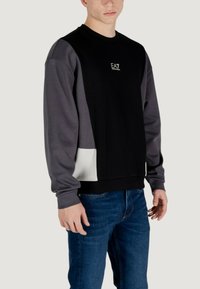 Felpa nera e grigia con maniche lunghe, caratterizzata da un design a blocchi di colore e logo sul petto, realizzata in tessuto morbido con una vestibilità comoda.