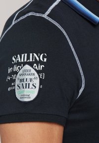 Granatowa koszulka polo z kontrastującymi białymi przeszyciami, naszywką z logo "Blue Sails" oraz napisem "Sailing in light Air" na ramieniu.