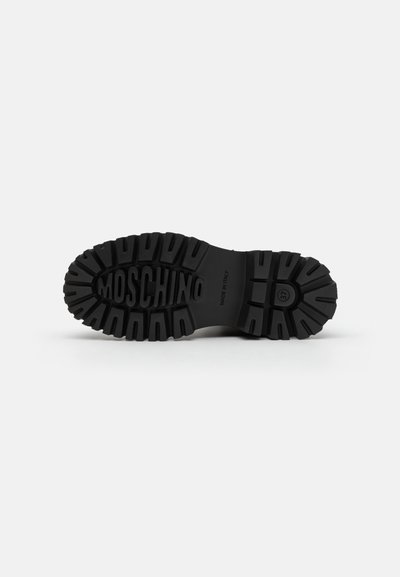 Crni gumeni potplat s dubokim uzorkom, s urezanim imenom brenda "Moschino" i oznakom "Made in Italy." Ukupna površina je glatka.