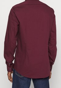 Burgundowa koszula z długim rękawem, o standardowym kołnierzyku, z guzikami przy mankietach i prostym dołem, wykonana z gładkiego materiału. Widok z tyłu ukazuje dopasowany krój.