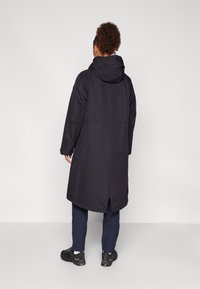 Palton lung negru, impermeabil, cu glugă, având o croială de lungime medie, slituri laterale și o textură netedă. Purtați cu pantaloni bleumarin și încălțăminte neagră.