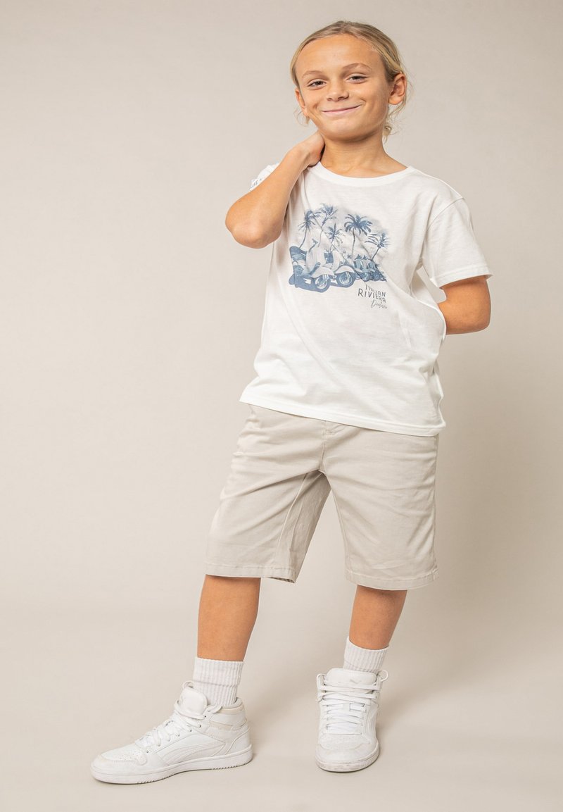 Garçon aux cheveux clairs portant un t-shirt blanc à motifs, un short beige, des chaussettes blanches et des baskets blanches, debout avec la main sur la nuque et souriant.