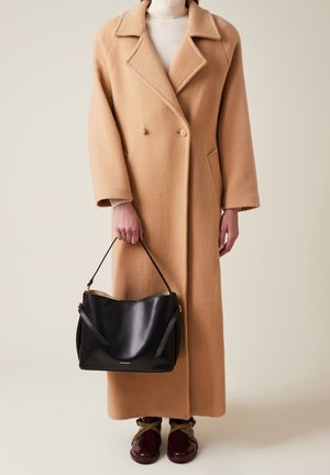 Cappotto in lana cammello con design doppiopetto, revers oversize e tasche laterali. Borsa a mano in pelle nera con finitura liscia e manici a spalla.