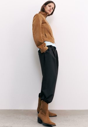 Nő fehér falnak dőlve, barna hasított bőr dzsekit, fekete laza nadrágot és hozzáillő barna hasított bőr bokacsizmát visel.