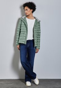 Ljust grön pufferjacka med huva i grått, två framfickor, över en vit topp och mörkblåjeans, ihop med vita sneakers.