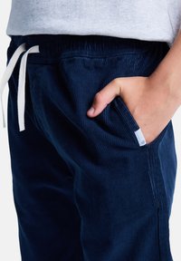 Morskie sztruksowe spodnie dresowe o teksturowanej powierzchni, z elastyczną talią i białym sznurkiem. Widoczna boczna kieszeń z małą metką z logo.