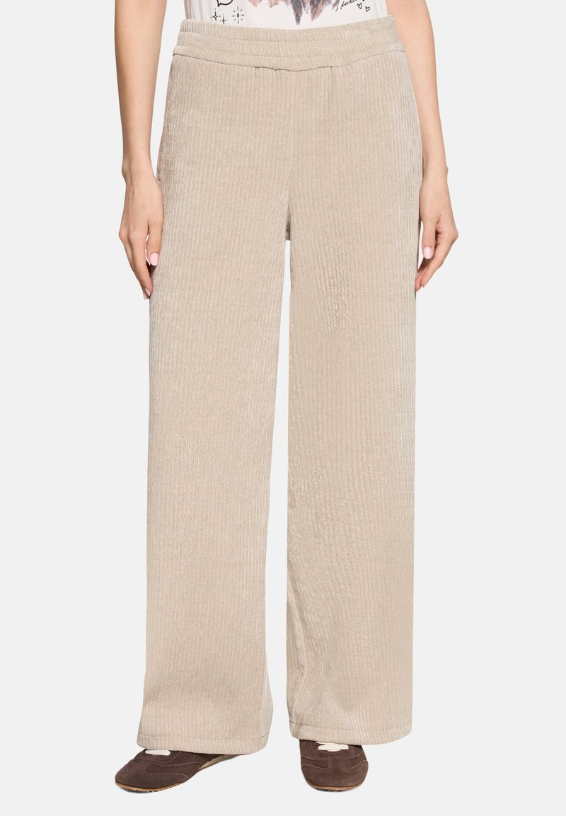 Pantalon beige à jambes larges à côtes, porté avec des chaussures marron et un haut blanc rentré, visible de la taille aux pieds sur un fond uni.