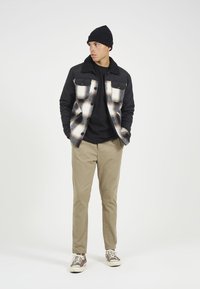 Podložena črna jakna s karirastim vzorcem v odtenkih smetane in rjave, črna kapa, črna majica, peščene hlače in rjavi športni čevlji.