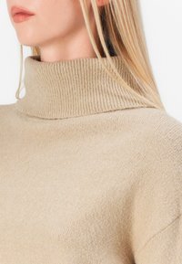 Beige polokragejumper med ribbad krage, tillverkad av mjukt, strukturerat stickat material; åtsittande design som visar en ren, minimalistisk look.