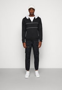 Felpa nera con accenti bianchi, colletto con zip e foderatura grigia nel cappuccio, abbinata a pantaloni cargo neri con tasche con zip e polsini elasticizzati. Sneakers bianche.