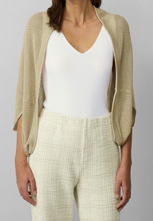 Femme portant un haut blanc à col en V, un pantalon beige texturé taille haute, et un châle en tricot beige ample sur les épaules.