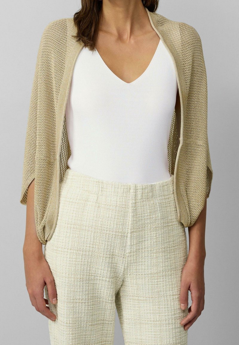 Femme portant un haut blanc à col en V, un pantalon beige texturé taille haute, et un châle en tricot beige ample sur les épaules.