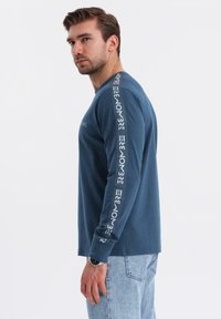 Muž v modré košili s dlouhým rukávem, na které jsou bílé nápisy na rukávech a hrudi, v kombinaci s světle modrými džíny a hodinkami na zápěstí.