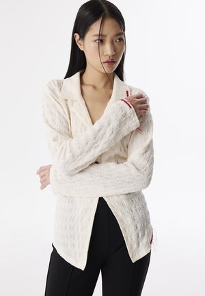 Off-white textured shirt with a collar, long sleeves, and a slightly curved hem; features red stitching accents on cuffs.