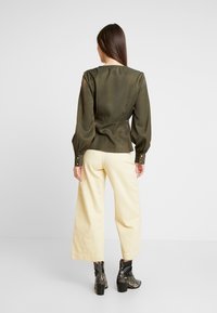 Olivengrüne Satinbluse mit Puffärmeln und Knopfdetails, kombiniert mit cremefarbenen, weiten Hosen und strukturierten, schwarzen Stiefeletten.