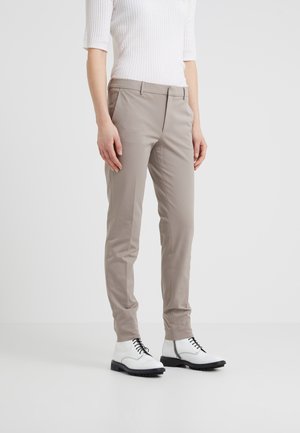 Person iført beige slim-fit bukser, hvid ribbet kortærmet top og hvide snørebåndsstøvler, der står på en ensfarvet hvid gulv.