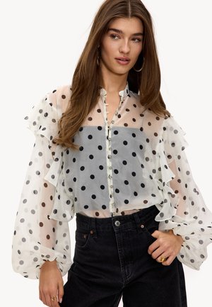 Jeune femme portant une blouse blanche transparente à pois noirs avec des manches à volants, associée à un jean noir taille haute et des bagues dorées.